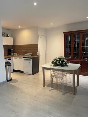Eine moderne Küche mit weißen Schränken, einem Geschirrspüler und einer Kaffeemaschine. Es gibt eine Essecke mit Tisch und Stühlen sowie einen Schrank mit Glastüren.