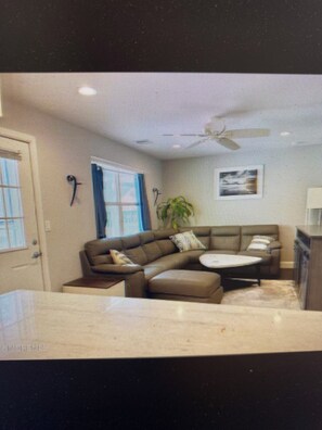 A modern living room with a sofa, a coffee table, and a small table. There is a window with blinds, a ceiling fan, and a plant.