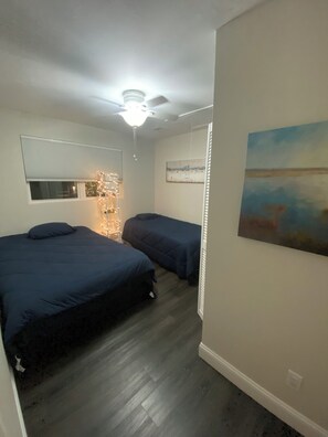 A bedroom with two beds, a ceiling fan, a window with blinds, and a painting on the wall.