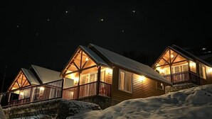 A row of wooden cabins with illuminated windows and balconies, set against a snowy landscape at night. - The Auli Meadows (Joshimath)