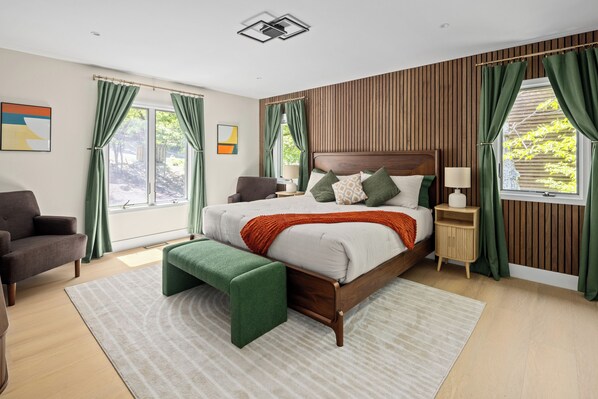 Quarto com cabeceira de madeira, um banco verde, cama com almofadas, mesa de cabeceira e uma janela com cortinas.
