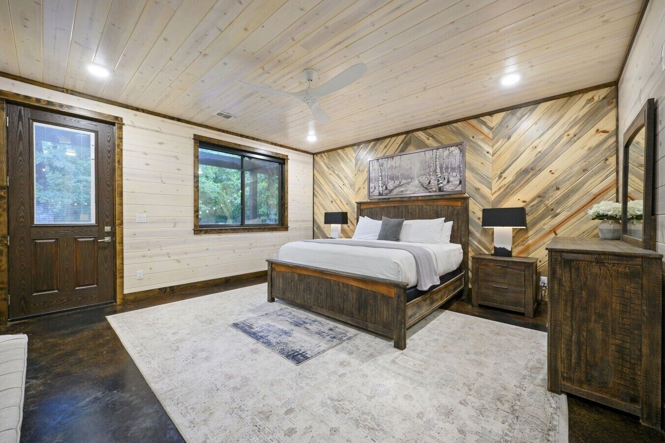 Quarto com teto de madeira, cama com roupa de cama branca, mesa de cabeceira, cômoda e uma janela grande.