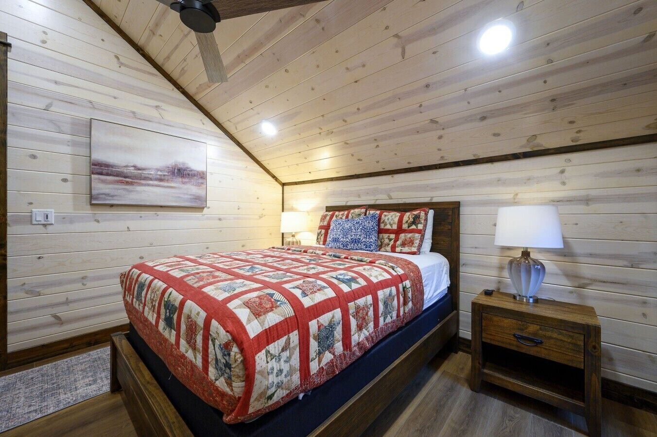Une chambre avec un plafond en bois, un lit recouvert d’une courtepointe, une table de chevet avec une lampe et un tableau accroché au mur.