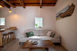 A cozy living area with a wooden ceiling, a sofa with cushions, a coffee table with a decorative item, a window with a view, and a painting on the wall. - Yiayia's House (Alonnisos)