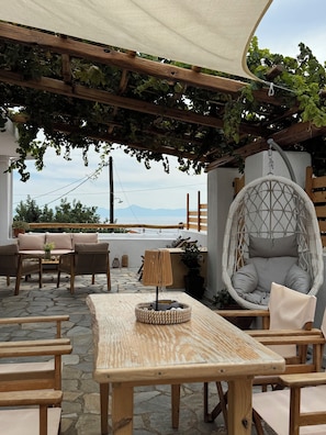 An outdoor seating area with a wooden table, wicker chairs, a hanging chair, and a view of the mountains. - Yiayia's House (Alonnisos)