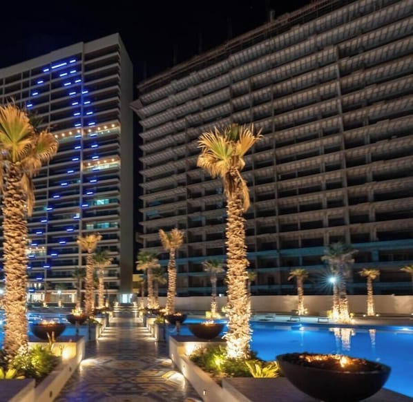 Un hôtel doté d’une piscine, de palmiers et offrant une vue nocturne sur les fenêtres illuminées du bâtiment.