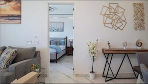 Una sala de estar moderna con un sofá, una mesa de centro con objetos decorativos y vistas a un dormitorio con cama y mesita de noche.