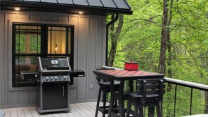 Een terras met een barbecue, een tafel en stoelen, omringd door een bos.