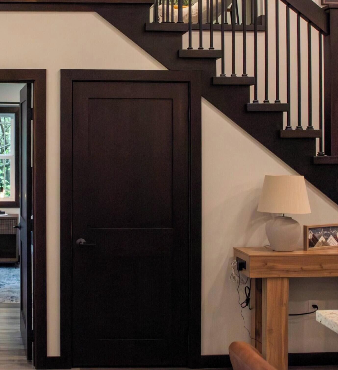 Une porte en bois sombre, un escalier avec des rampes noires et une table de chevet en bois surmontée d’une lampe.