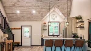 A kitchen with a wooden ceiling, a clock, bar stools, and a window.