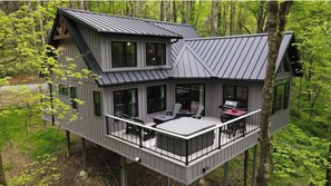 A two-story cabin with a balcony, large windows, and a metal roof, surrounded by forest.