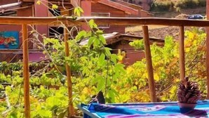 Un balcón de madera con una mesa y un cojín, con vistas a un paisaje rural con casas y árboles.
