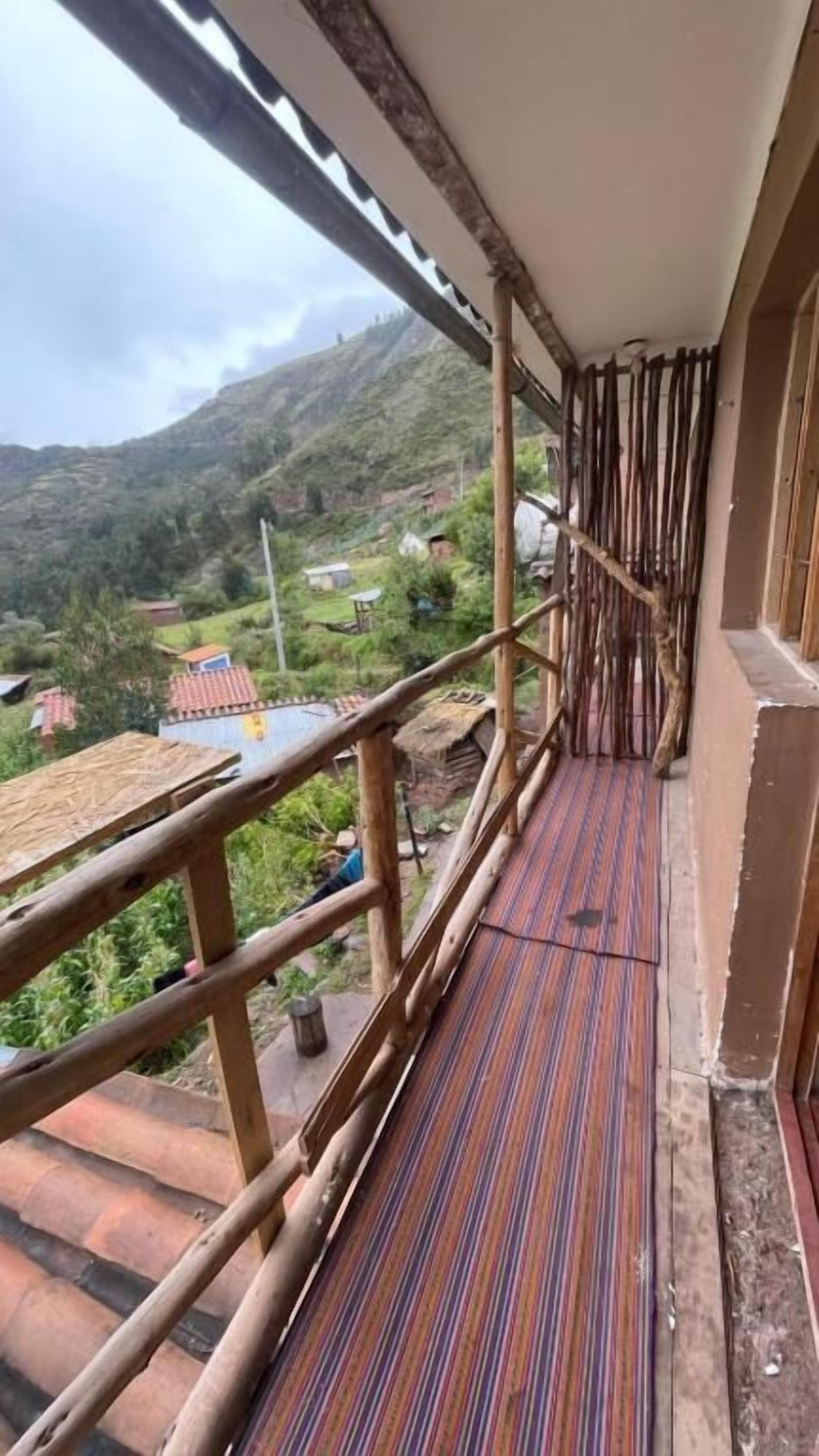Un balcon doté d’un plancher en bois et d’une rambarde en métal, offrant une vue sur une région montagneuse parsemée de maisons.