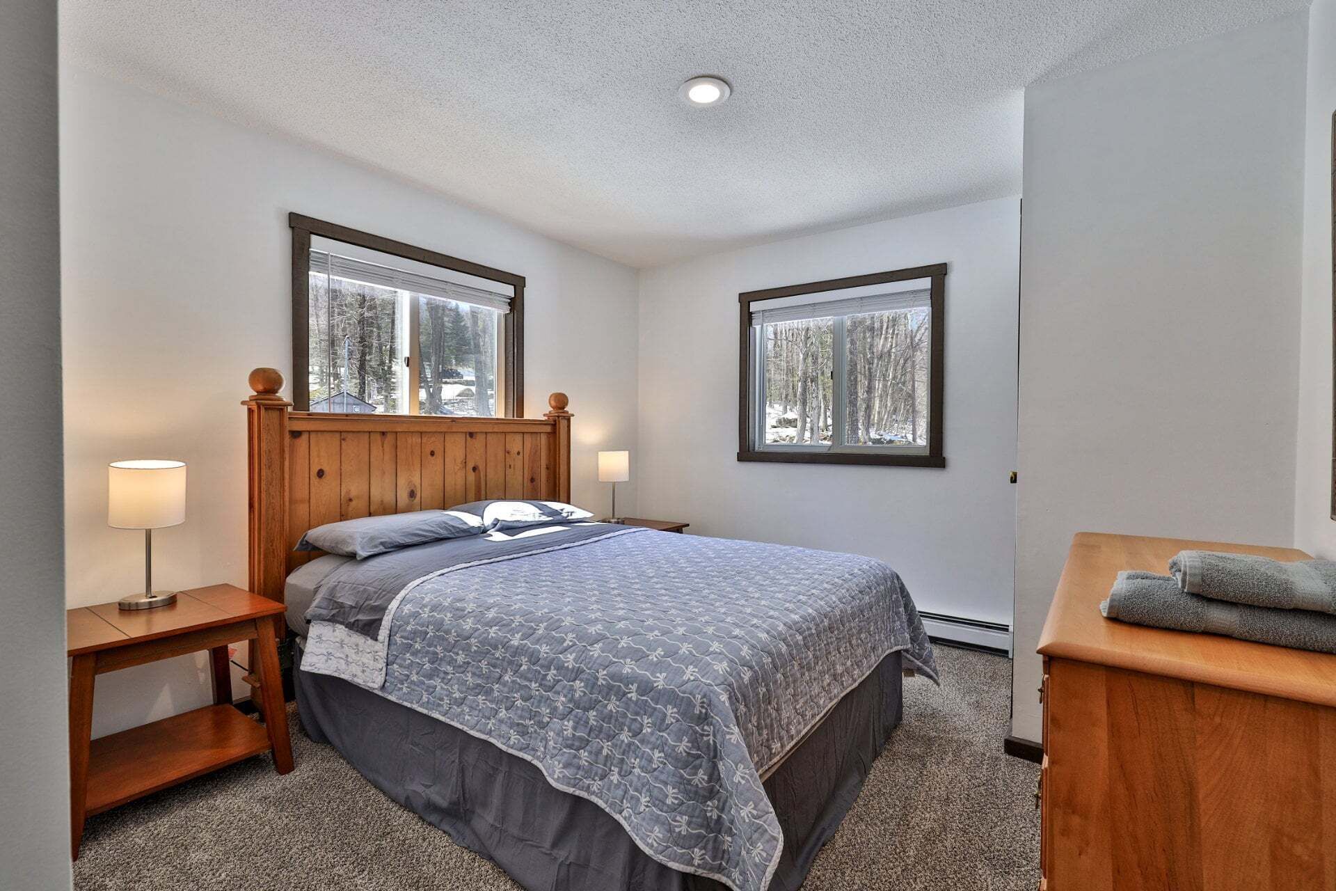 A bedroom with a wooden headboard, two bedside tables with lamps, a bed with a gray patterned bedspread, and two windows with a view of trees.