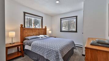 A bedroom with a wooden headboard, two bedside tables with lamps, a bed with a gray patterned bedspread, and two windows with a view of trees.