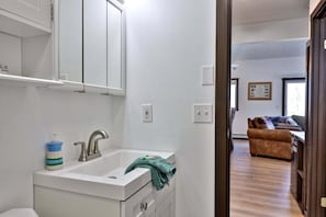 Ein Badezimmer mit Waschbecken, Spiegel und Schrank, ein Flur, der in einen Wohnbereich mit einer Couch und einem Fenster führt.