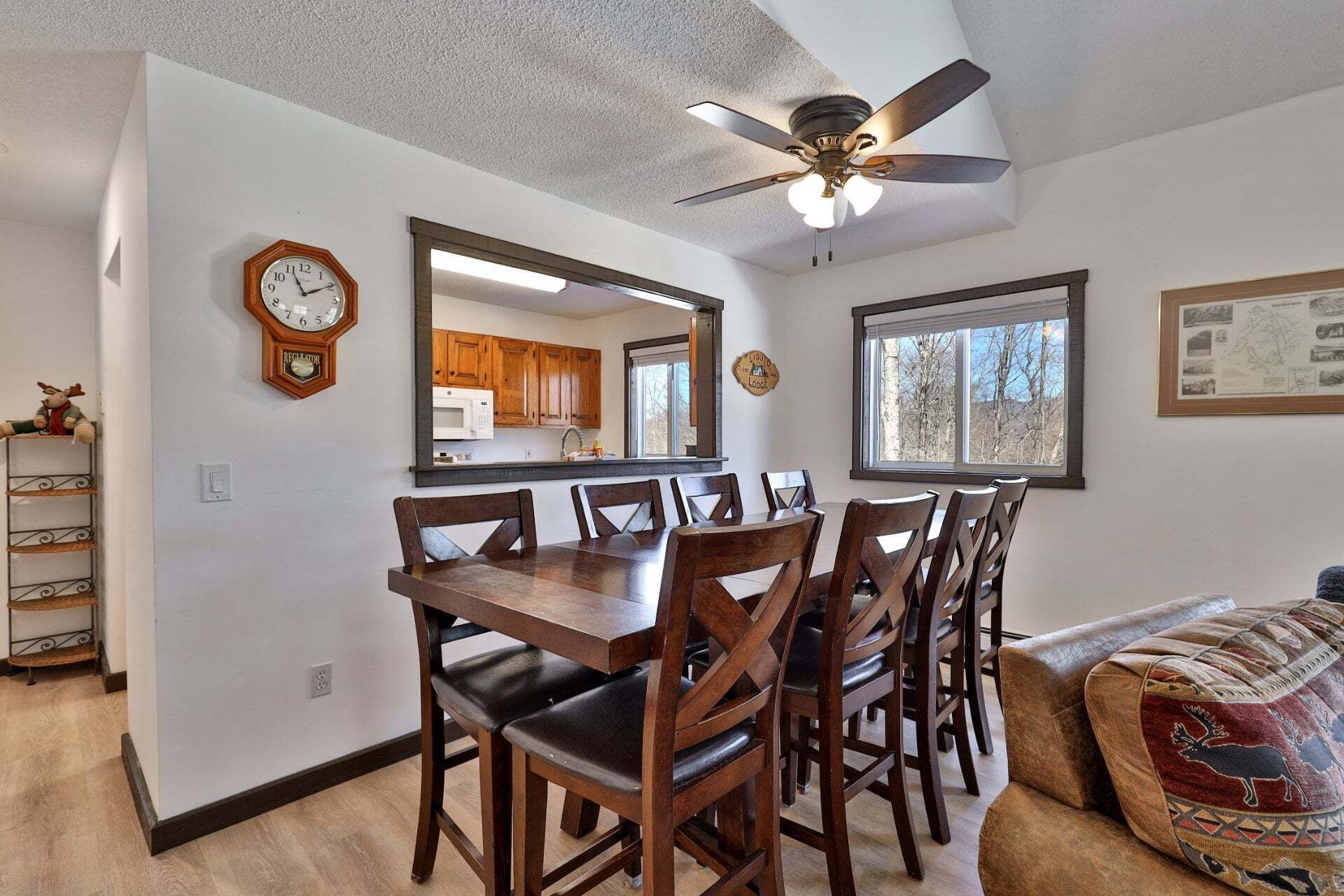Un coin repas avec une table et des chaises en bois, un ventilateur de plafond, une horloge, une fenêtre offrant une vue agréable et une cuisine visible à travers un miroir.