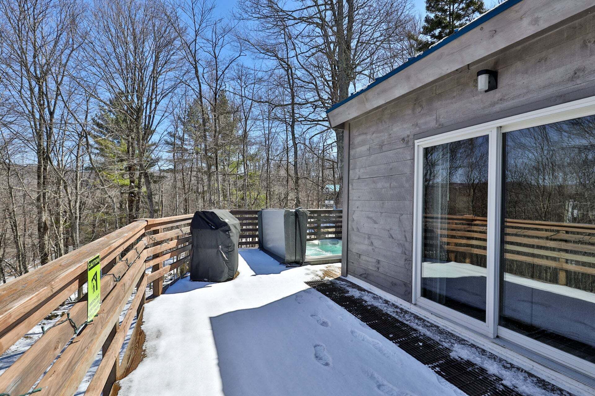 A wooden deck with a snow-covered ground, a large window, and a hot tub area.