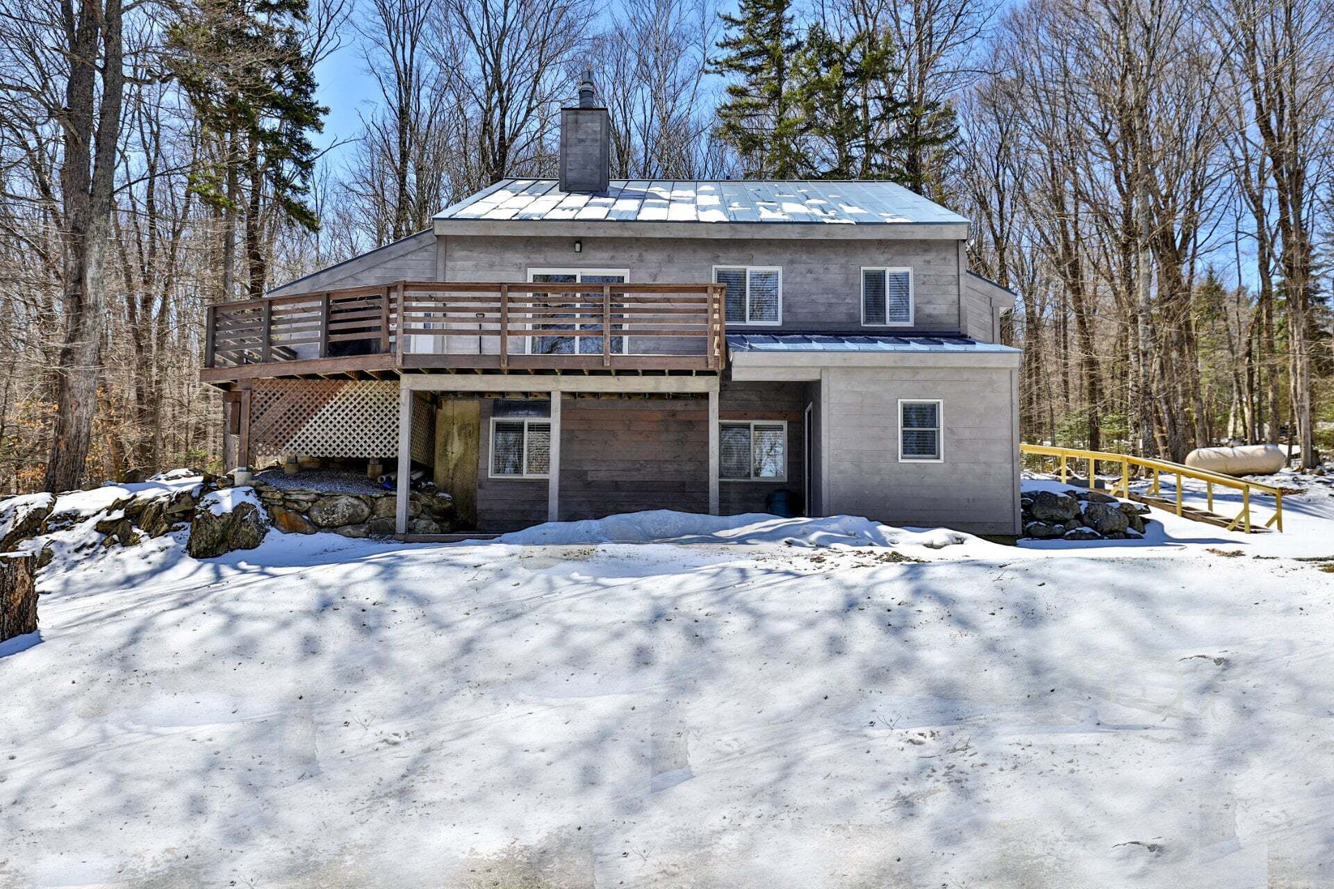 A two-story cabin with a wooden deck and a covered porch, surrounded by a snowy landscape and trees.