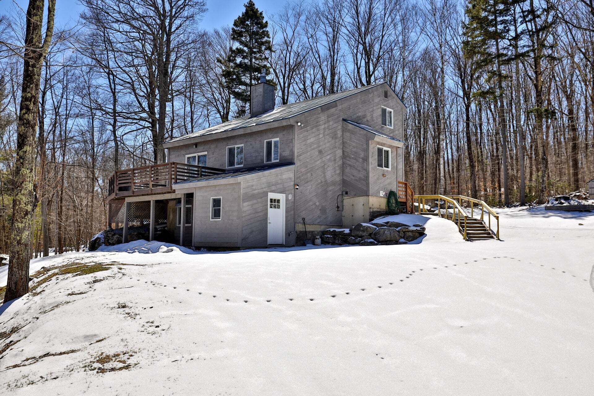 A two-story house with a deck, surrounded by a snowy landscape and trees.