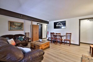 Ein möbliertes Wohnzimmer mit einer Couch, einem Couchtisch aus Holz und einem Esstisch mit Stühlen. An der Wand hängt ein Gemälde, und ein Schild mit der Aufschrift 'simple vacation rentals' ist sichtbar.