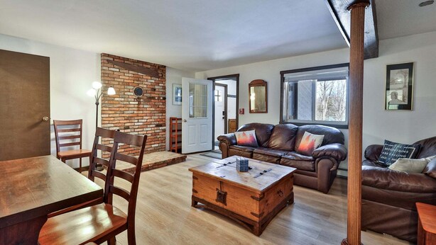 A living room with a wooden table, two chairs, a leather sofa, a coffee table, and a brick wall. - Liquid Lodge Lower: 2BR Duplex near Killington with 4 PM Checkout (Killington)