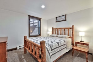 A bedroom with a wooden bed, a nightstand, a lamp, a window with blinds, and a wall-mounted light fixture. - Liquid Lodge Lower: 2BR Duplex near Killington with 4 PM Checkout (Killington)