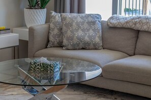 Ein modernes Wohnzimmer mit einem Glastisch, einem beigefarbenen Sofa und Kissen mit Muster.