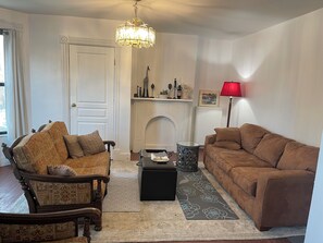 Een woonkamer met een open haard, twee banken, een kamerkleed met een patroon en een kroonluchter.