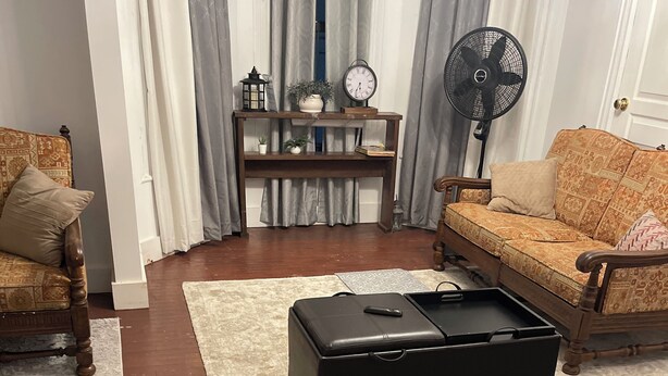 Olohuoneessa on puinen sohva, musta rahi, puinen sohvapöytä, pieni pöytä koriste-esineineen ja lattiatuuletin.