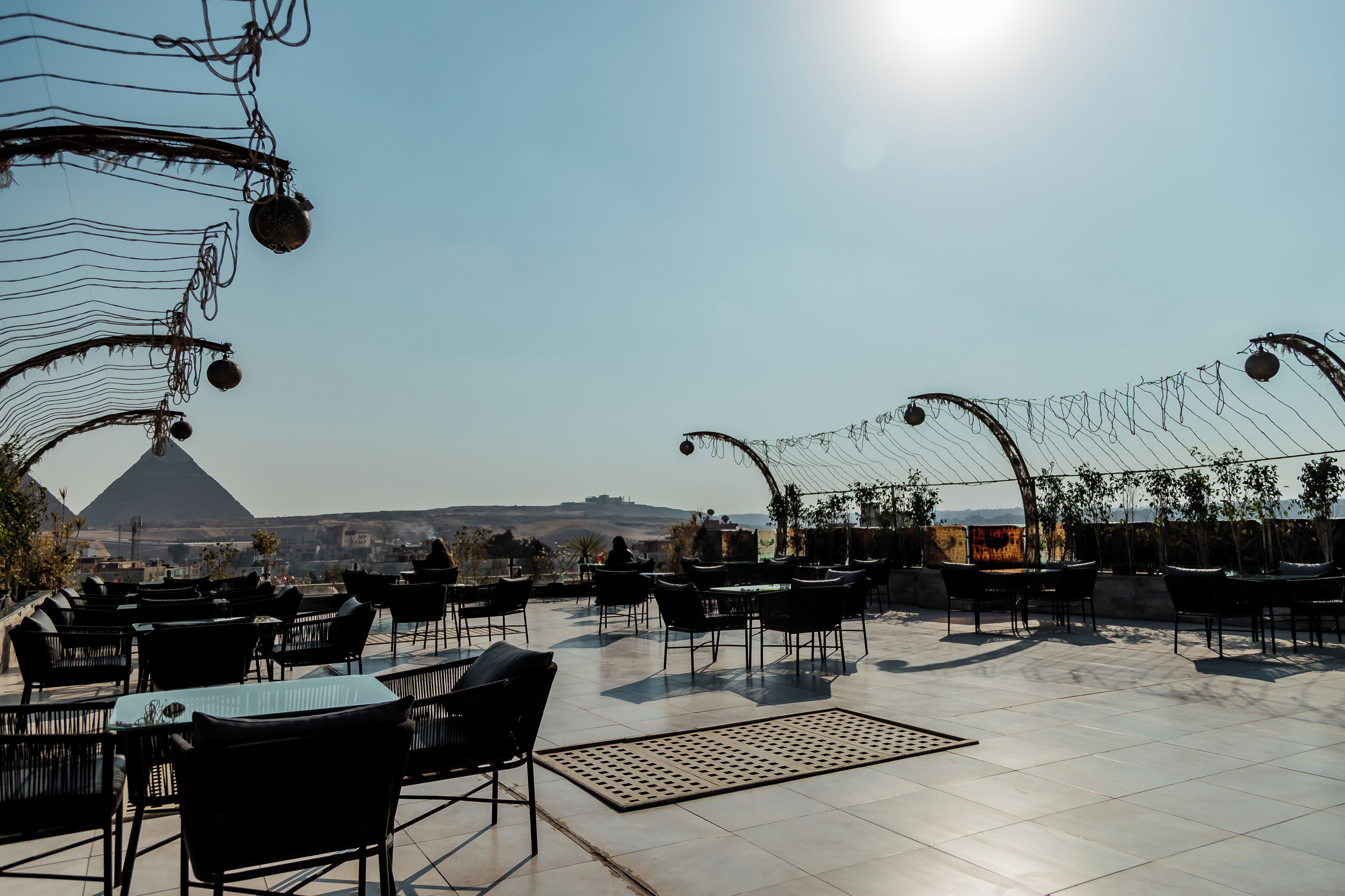An outdoor seating area with tables and chairs, a clear sky, and a distant view of a pyramid.