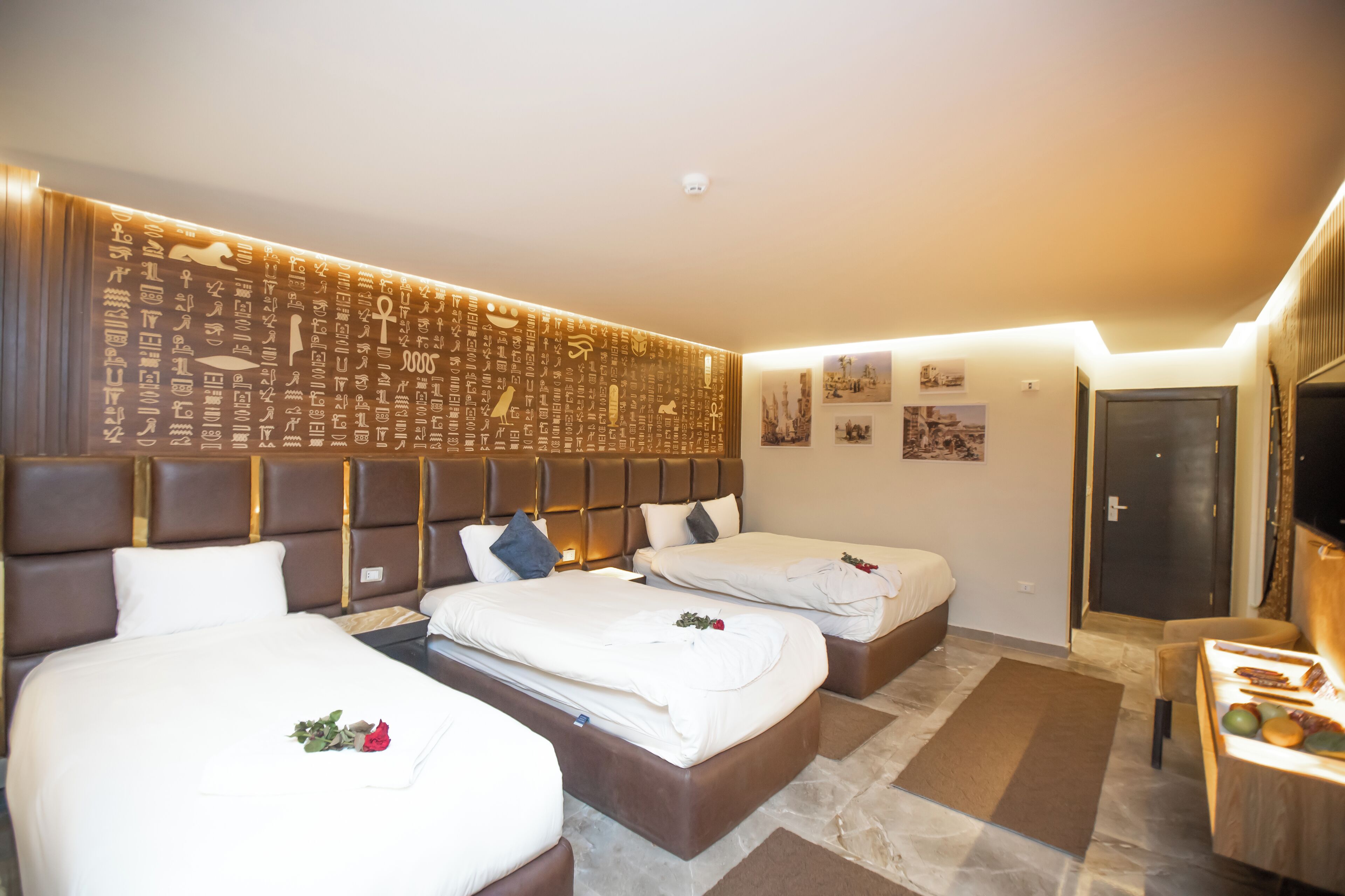 A hotel room with three beds, a decorative wall, a flat-screen TV, and a small table with fruits.