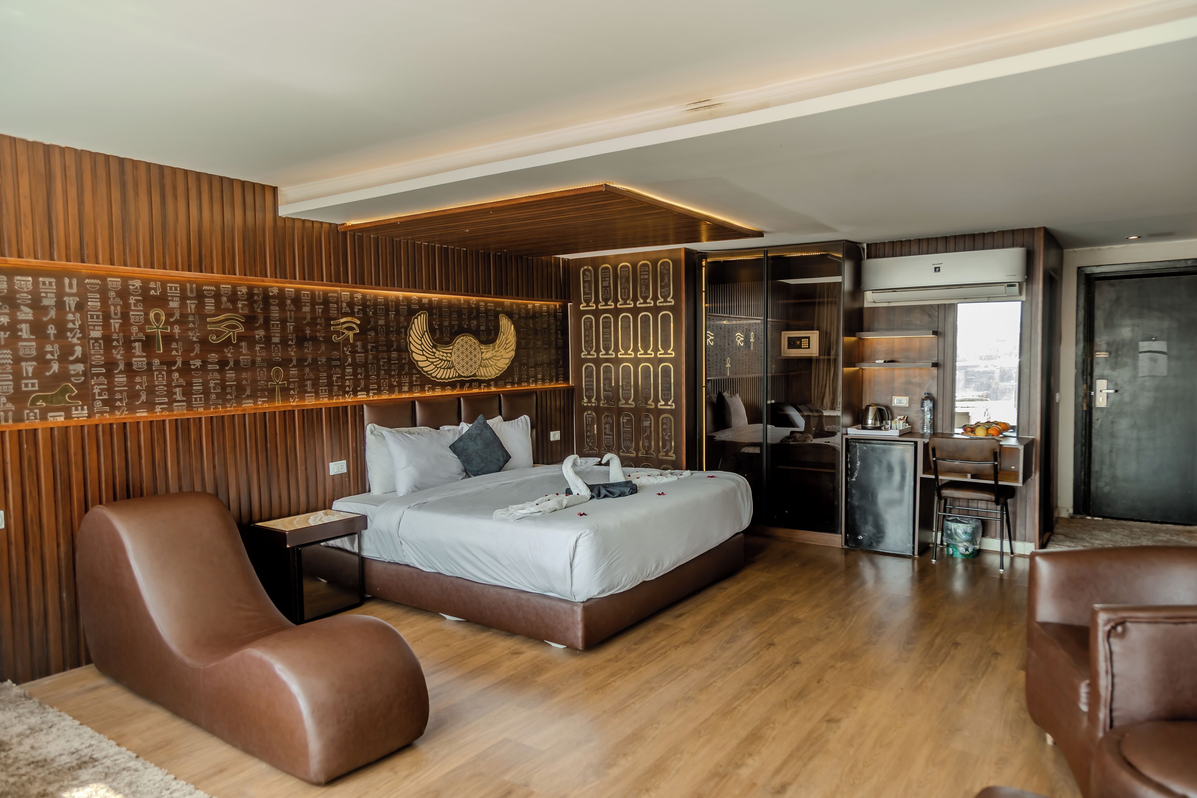 Una habitación de hotel moderna con una pared de acento de madera, una cama con cabecero decorativo, un área de descanso con un sillón de cuero marrón y una kitchenette.