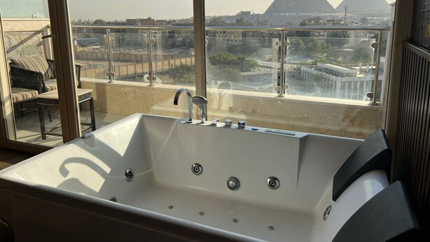 A modern bathroom with a bathtub, a glass-enclosed balcony, and a view of the pyramids. - Hotel Comfort Giza (Giza)