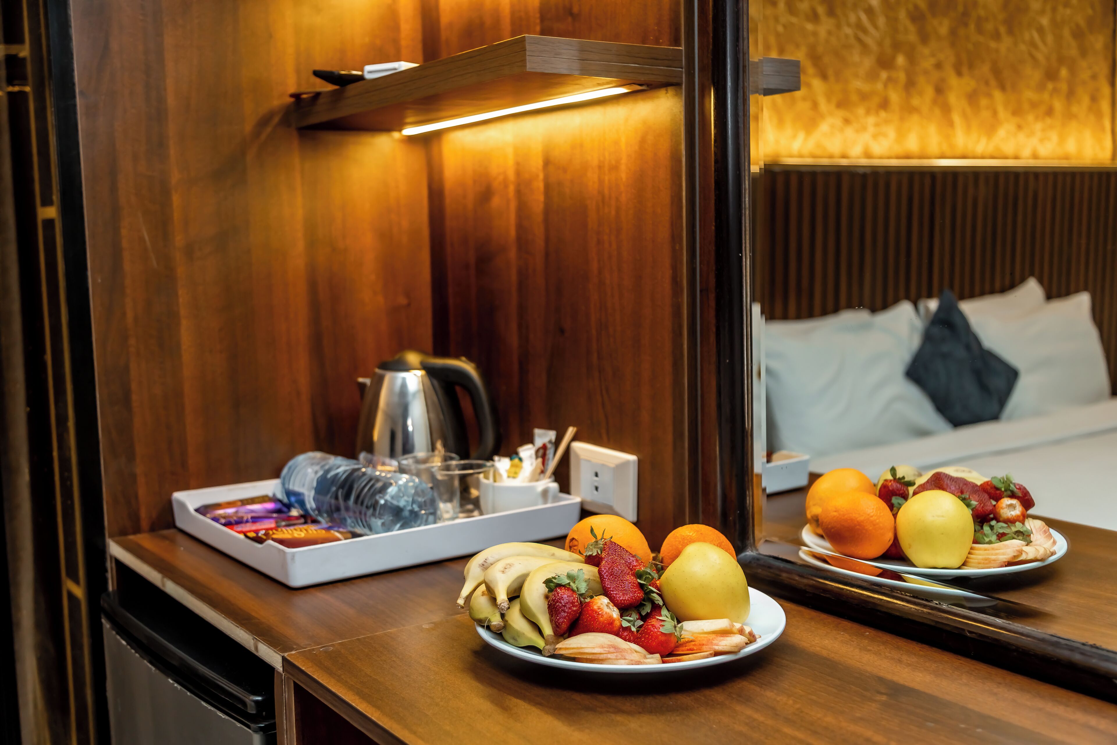 Ein Hotelzimmer mit einer Holzwand, einem Bett mit weißer Bettwäsche, einem Tablett mit Früchten und einem Wasserkocher, sowie einem Spiegel, der den Raum reflektiert.
