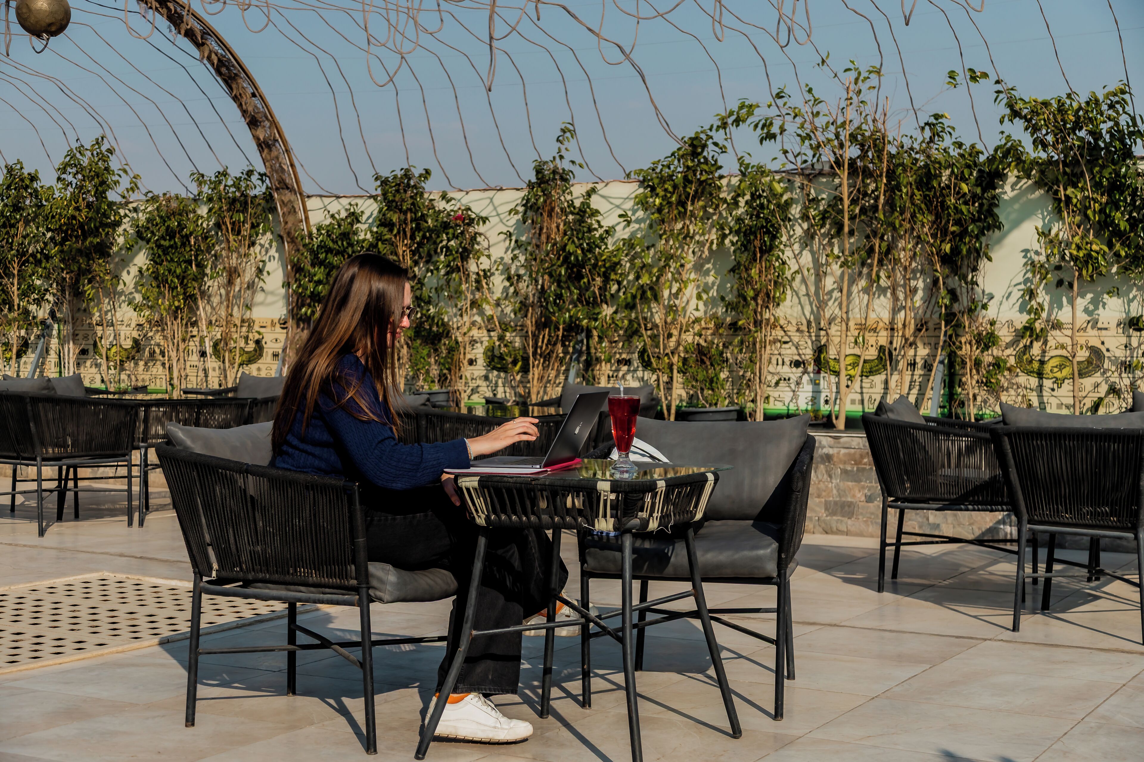 A person seated at a table with a laptop, surrounded by outdoor chairs and tables.