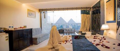 Une chambre d’hôtel équipée d’un lit, d’un bureau, d’une chaise, d’un canapé, d’un mini-bar et offrant une vue sur les pyramides.