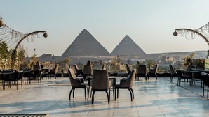 An outdoor seating area with tables and chairs, a view of the pyramids, and a clear sky. - Hotel Comfort Giza (Giza)