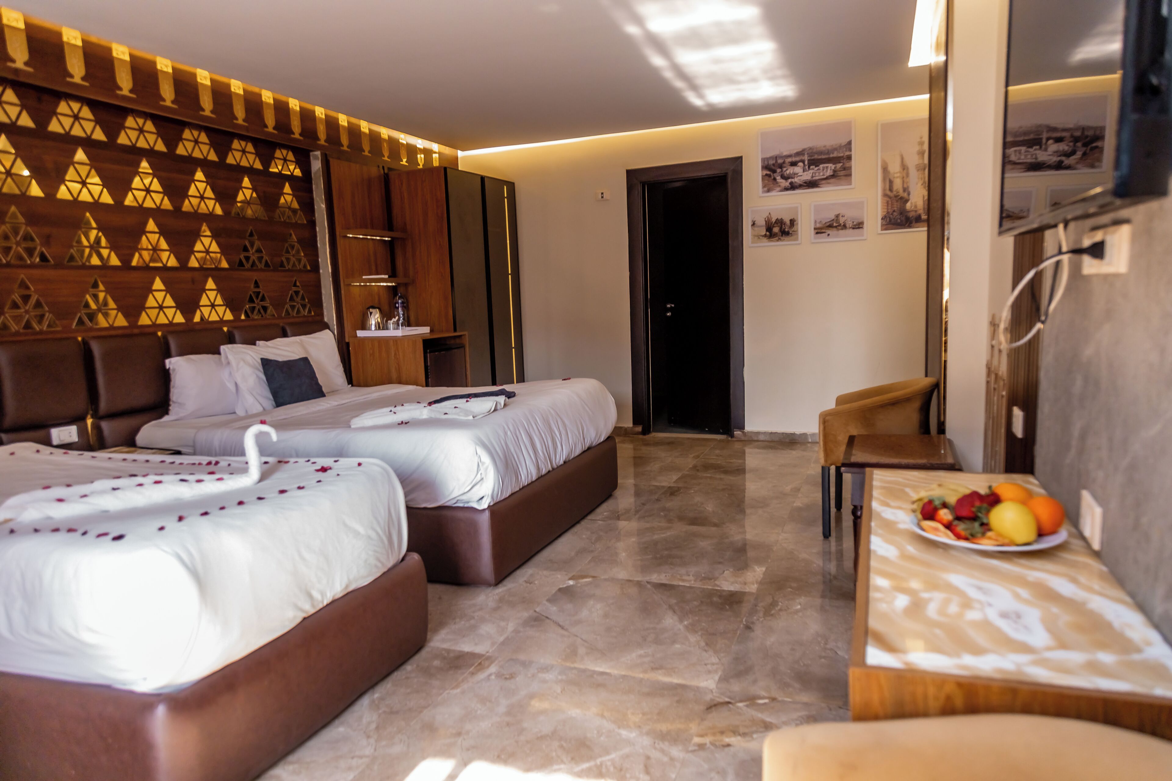 Habitación de hotel con dos camas, una mesa de comedor con fruta, cabecera de madera y un panel decorativo en la pared.