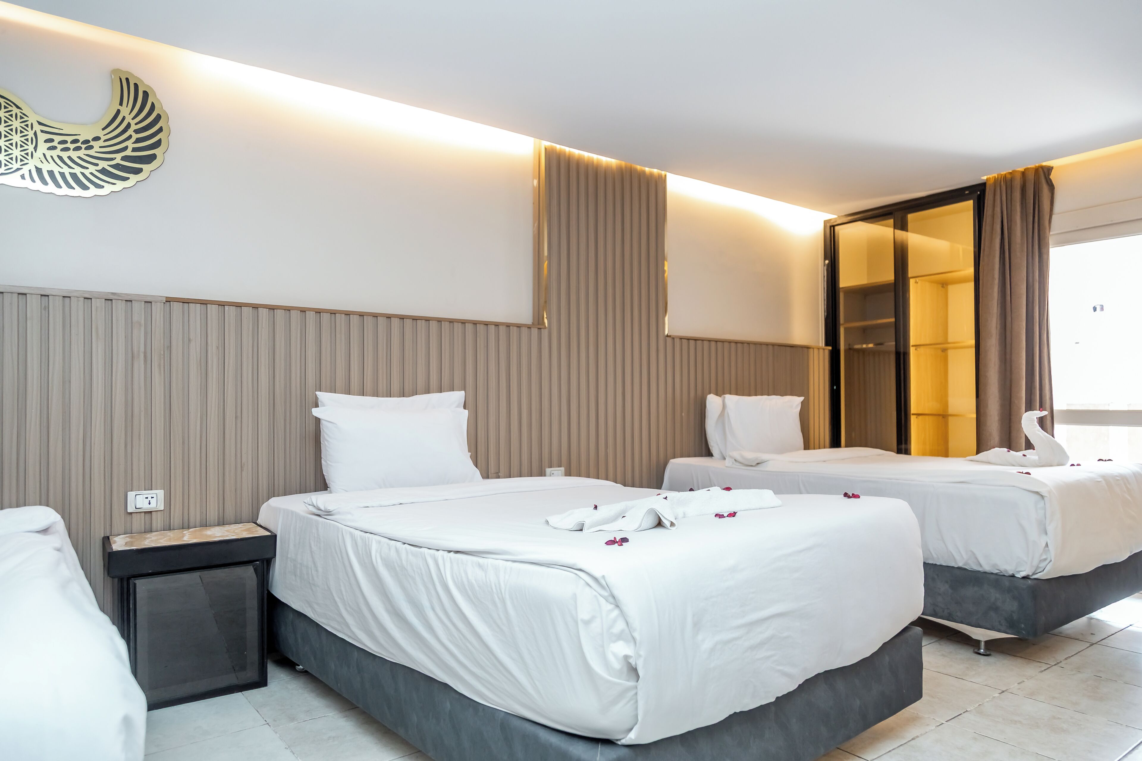 A hotel room with two beds, a wardrobe, a bedside table, and a wall-mounted light fixture.