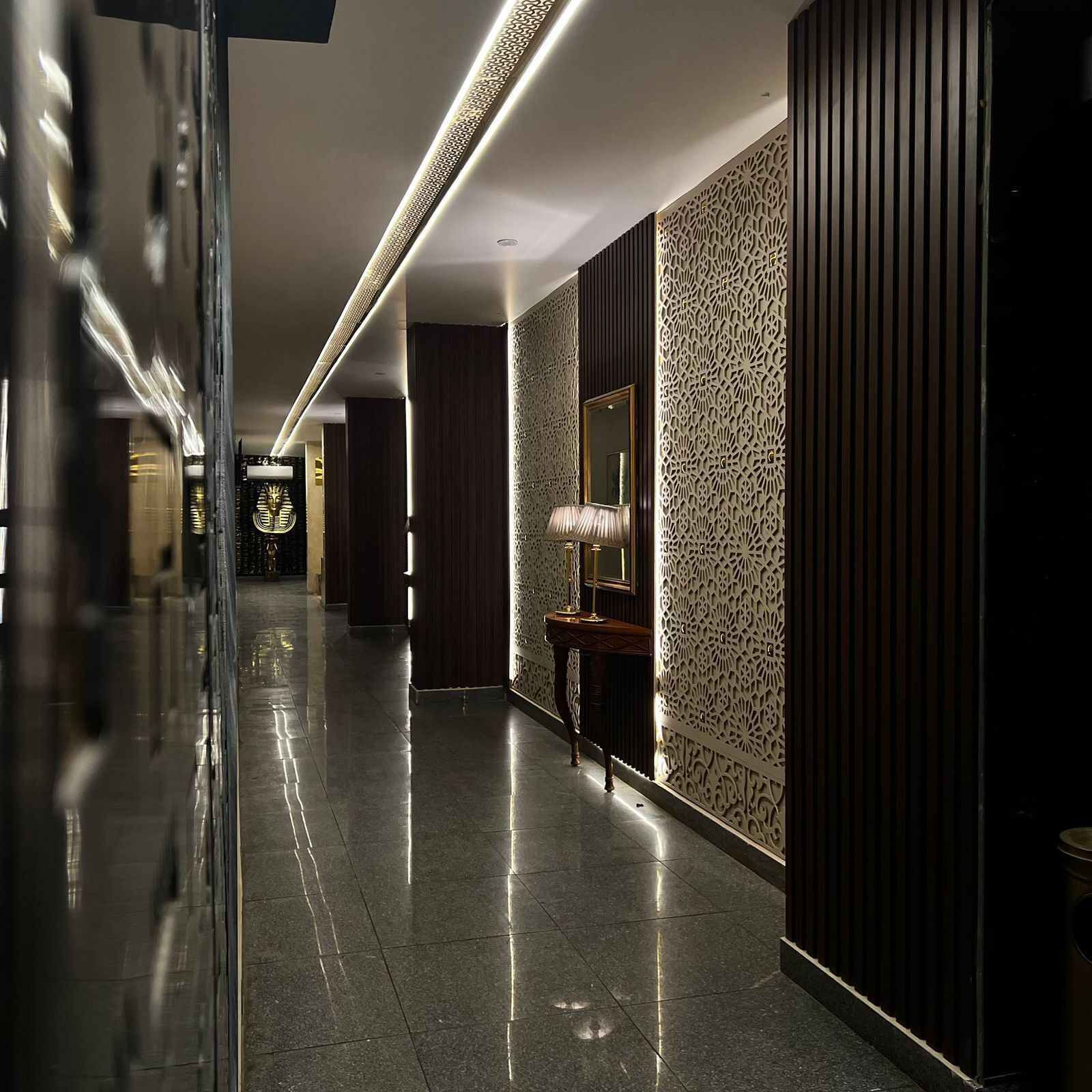 A corridor with reflective flooring, a decorative wall with intricate patterns, and a mirror on a console table.