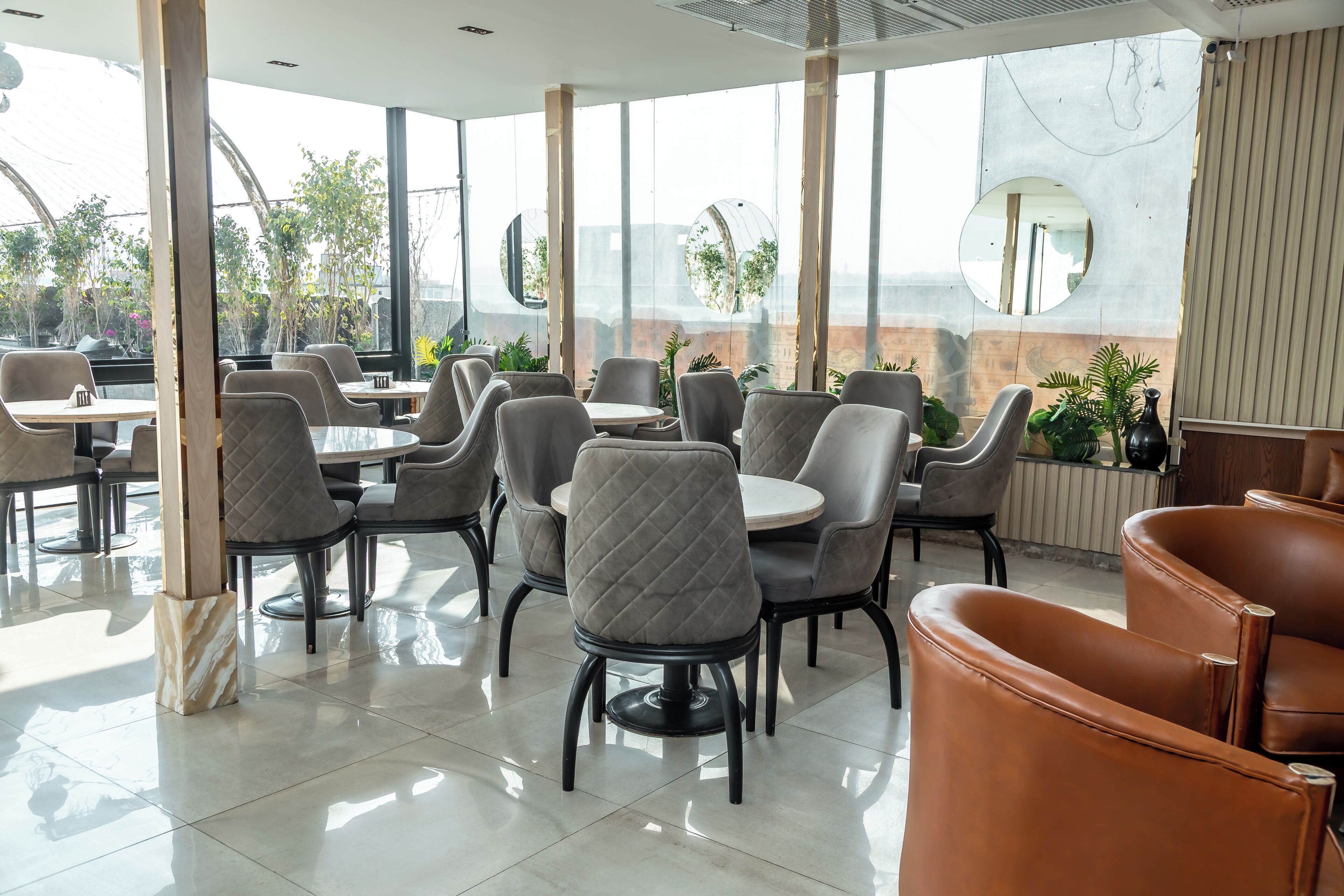 Een modern café met ronde tafels, stoelen met kussens, grote ramen en kamerplanten.