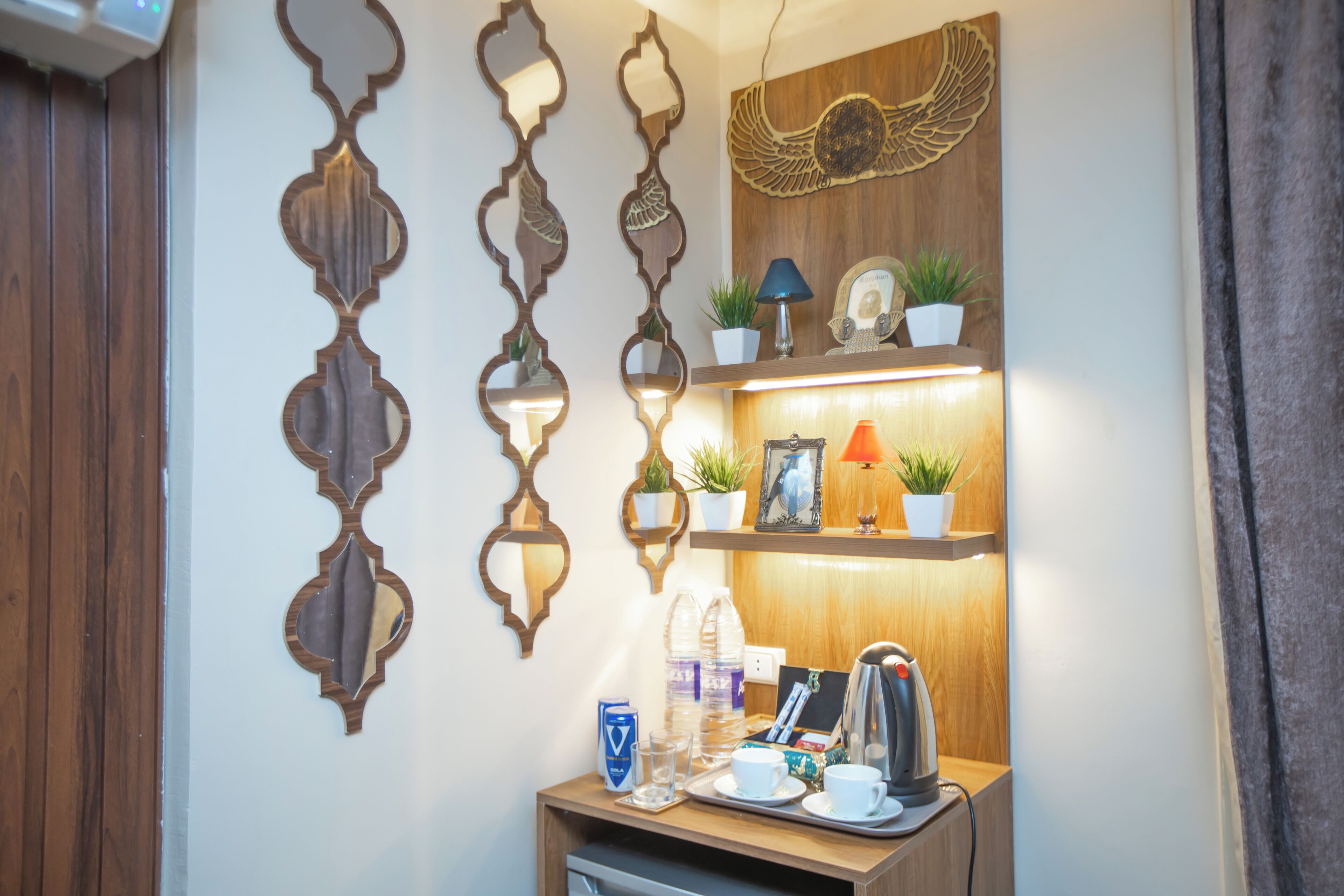 Un mostrador de recepción de hotel con espejos decorativos en la pared, un estante de madera con plantas, una lámpara y un hervidor.
