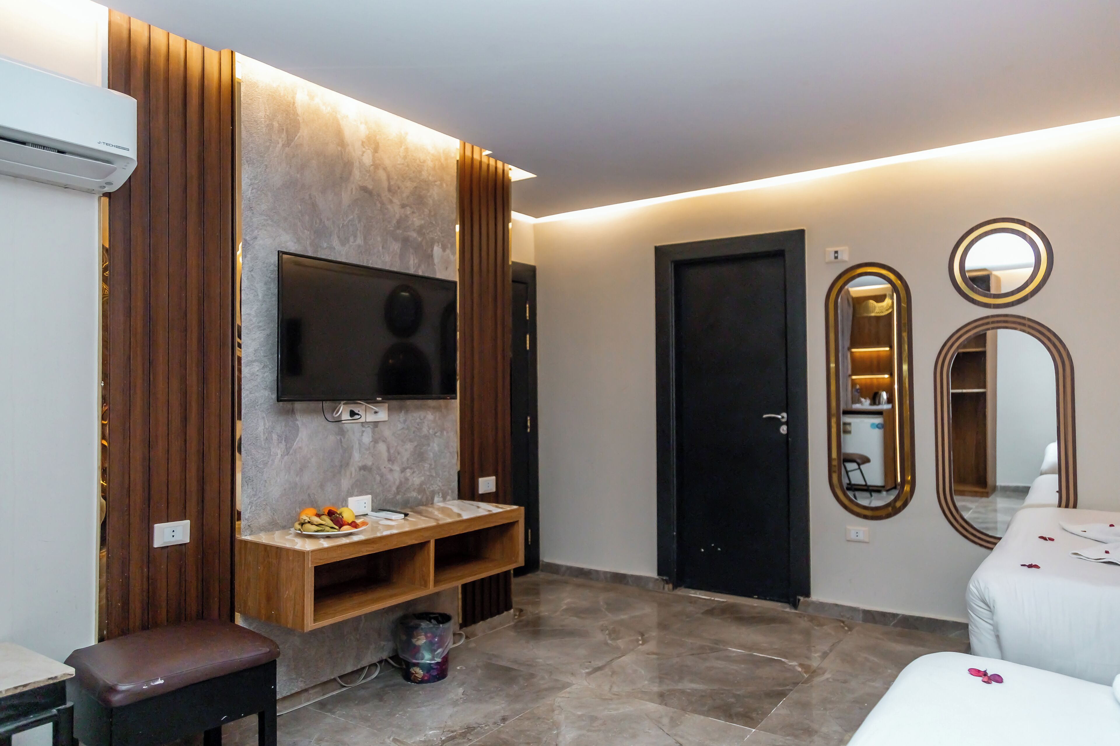 A modern hotel room with a flat-screen TV, wooden panel wall, a small wooden table, a bench, a bed, and two mirrors.