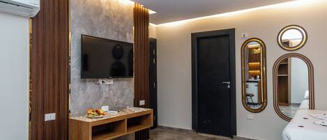 A modern hotel room with a flat-screen TV, wooden panel wall, a small wooden table, a bench, a bed, and two mirrors.