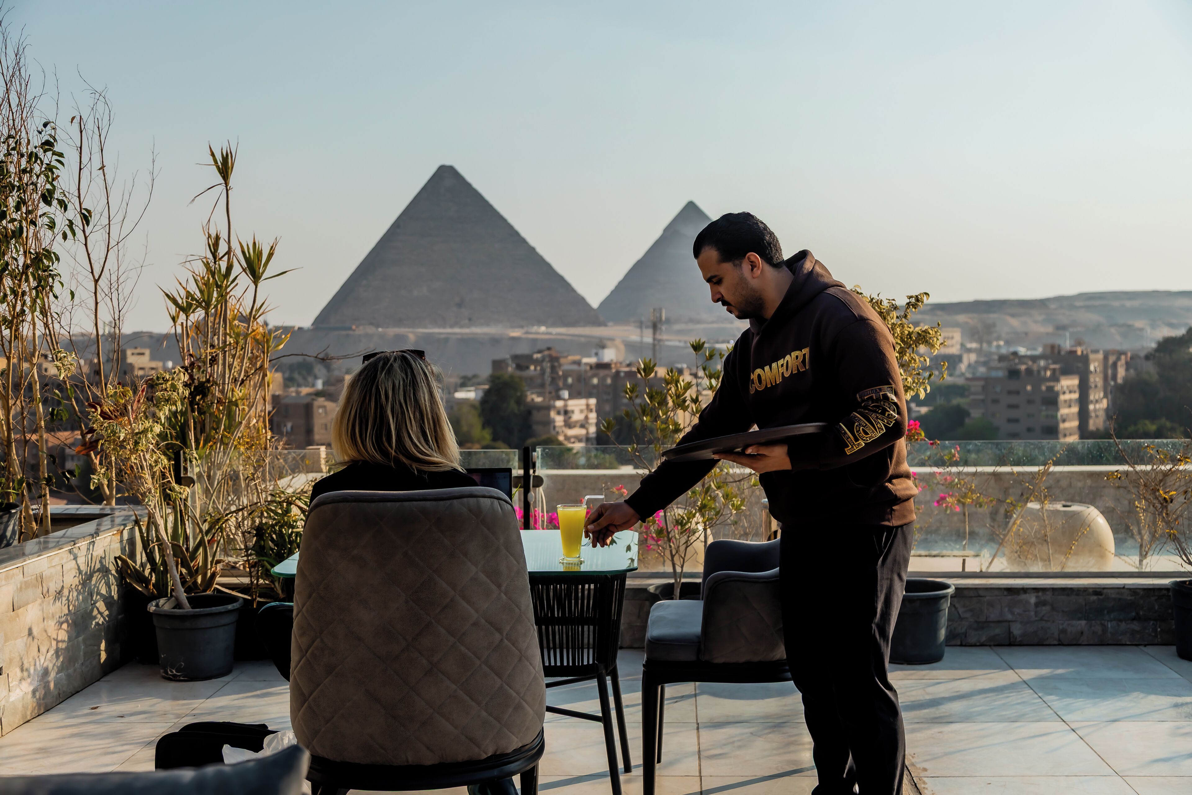 A person serving a drink to another person at a table with a view of pyramids.