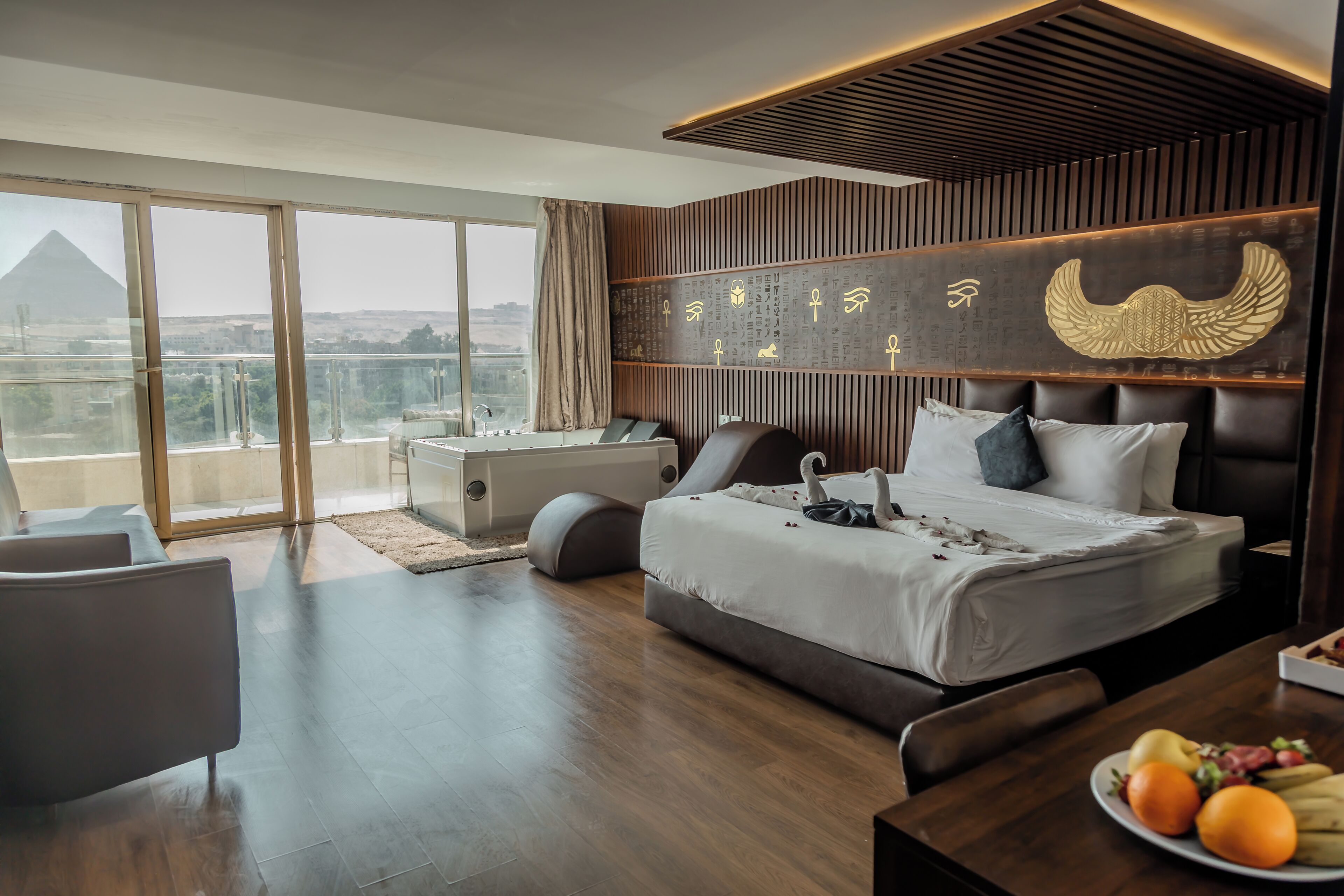 A modern hotel room with a large bed, a desk, a chair, and a dining table with fruit.