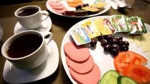 A breakfast setting with two cups of tea, sliced ham, tomatoes, cucumbers, olives, and cheese on a plate.