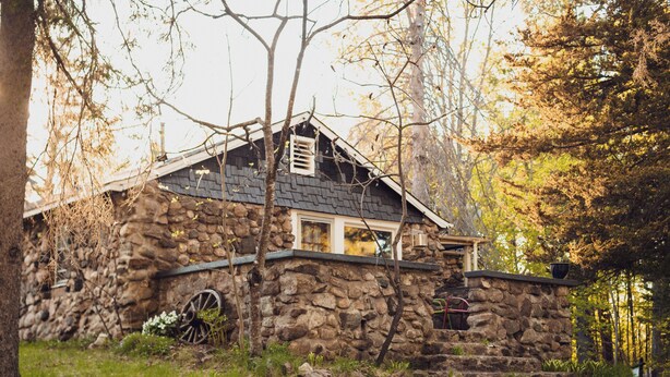 A stone house with a wooden door, a chimney, and a wheelbarrow in the yard.