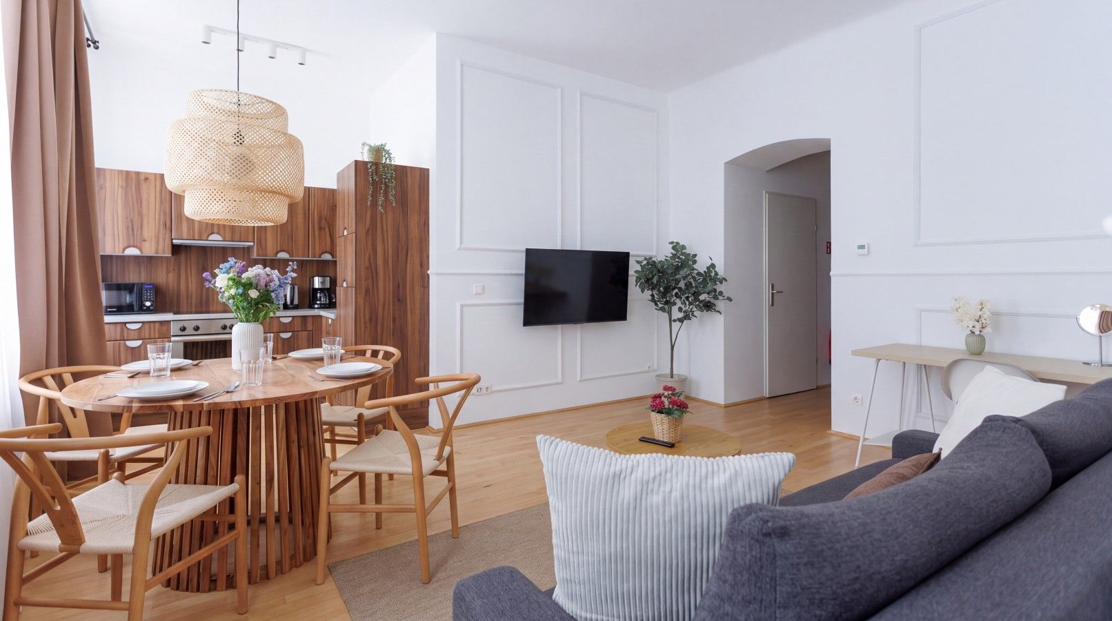 A modern living room with a grey sofa, a wooden dining table with chairs, a flat-screen TV mounted on the wall, and a kitchen area in the background.